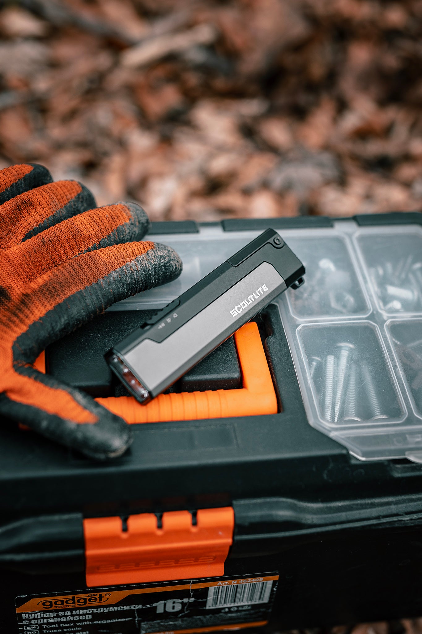 Orange and black glove holding a tool over a toolbox with a blurred natural background