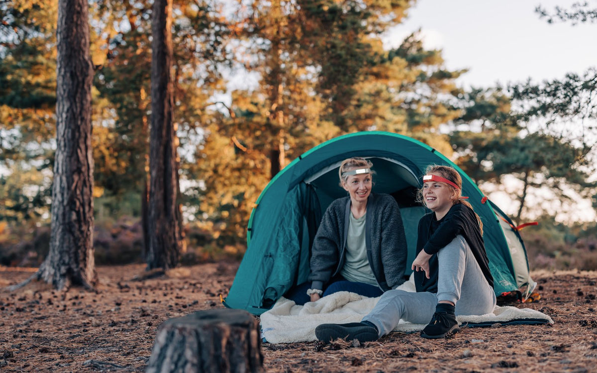 Two people outside a tent in the woods wearing lit Scoutlite headlamps